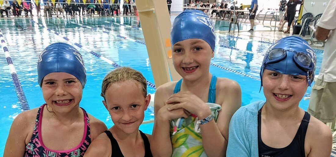 Four young Boundary Bay Bluebacks swimmers in blue caps smiling poolside at a summer swim meet in Delta, BC